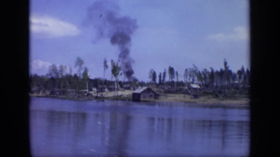 1971 Moscow-area river view with a dark industrial smoke plume rising behind a small cabin and sparse trees.