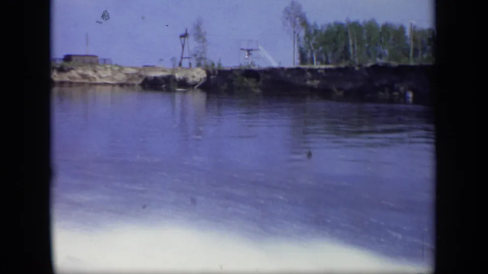 1971 Moscow 8mm frame of a low industrial riverbank with equipment, rough shoreline, and distant trees.
