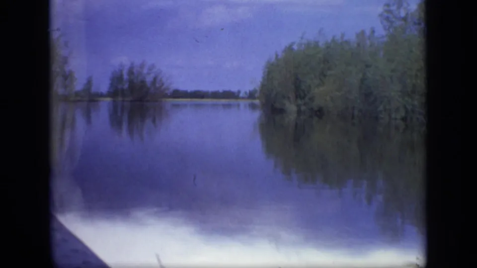 1971 8mm view near Moscow showing calm water, reed growth, distant tree line, reflections, and a pale blue sky.