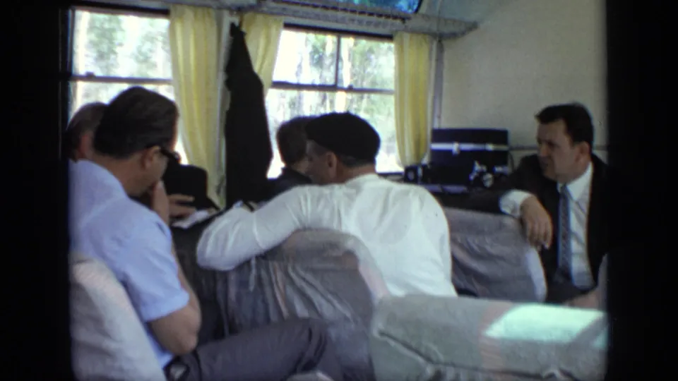 1971 8mm interior near Moscow showing seated adult men in shirts, suit jacket, tie, and cap beside curtained windows looking outward.