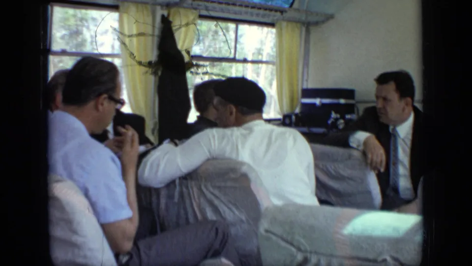 1971 Moscow-area home-movie frame of adult men talking inside a boat cabin with curtains, windows, and water outside.