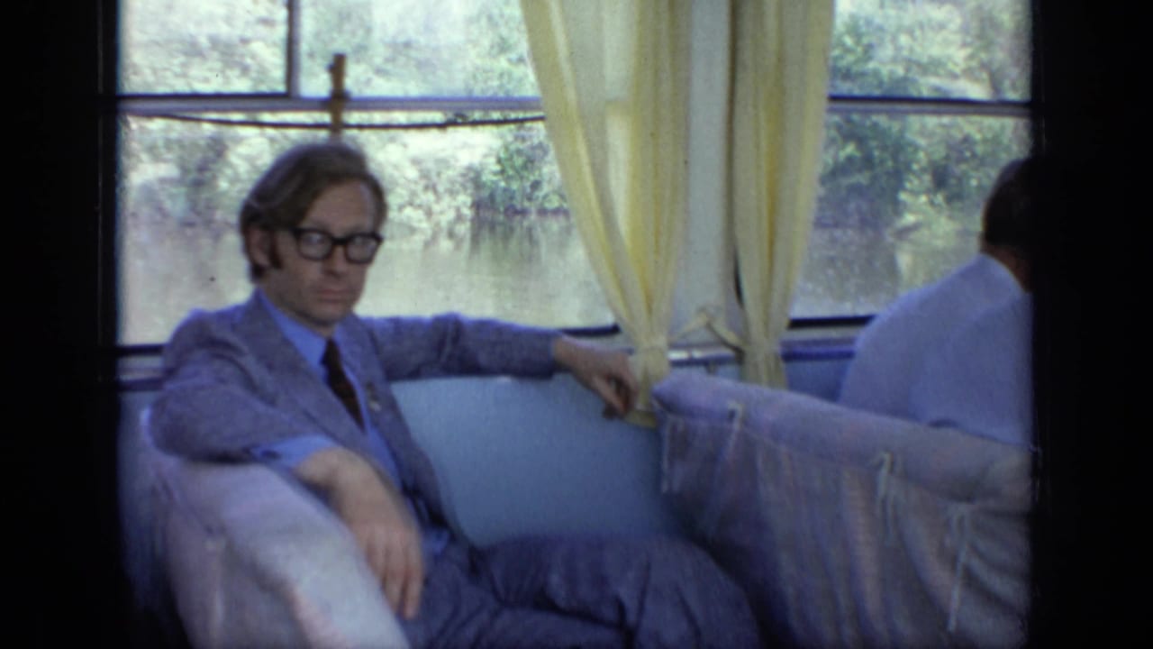 Man in glasses and a light suit seated by a boat window with yellow curtains.