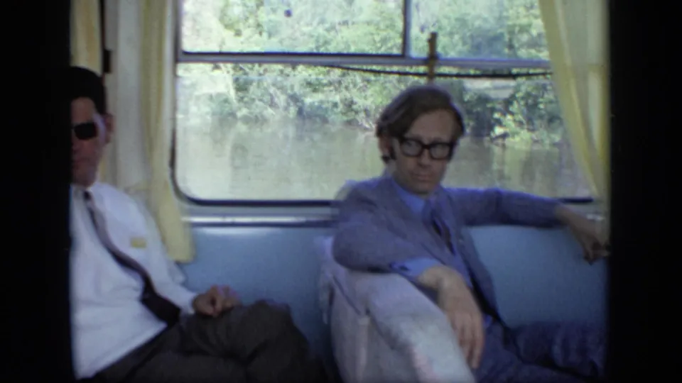 Two adult men sit in a boat cabin, with water and greenery outside the window in the 1971 Moscow-area home movie.