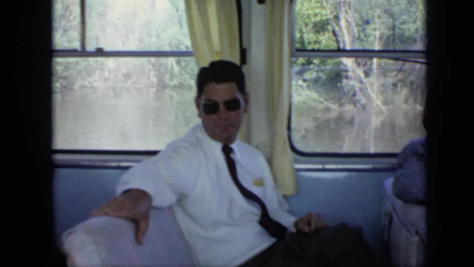 A man in sunglasses, white short-sleeve shirt, and tie sits by a boat-cabin window in the 1971 Moscow-area footage.