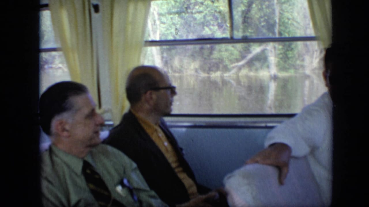 Several men in ties sit side by side near boat windows with trees outside.