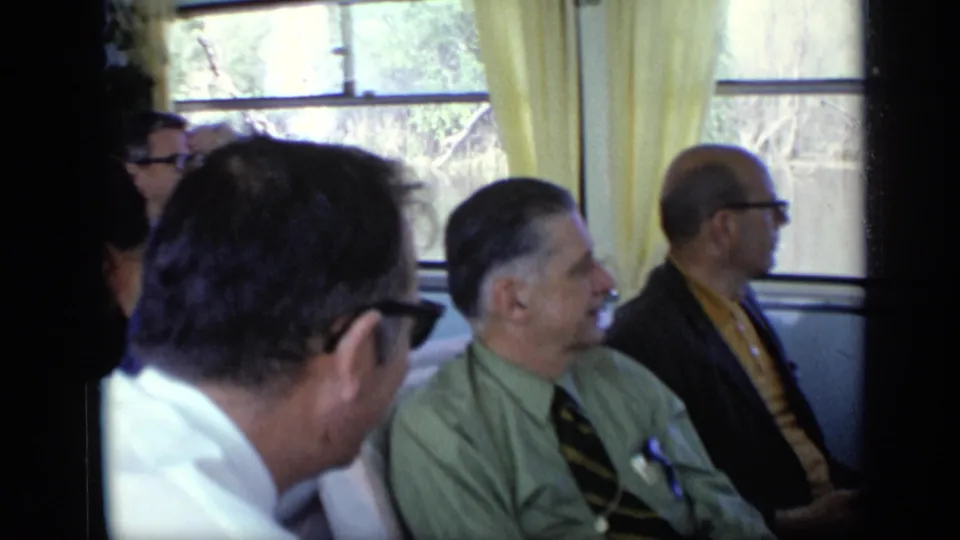 1971 home-movie frame showing suited passengers in a boat cabin, including a man in a green shirt and others seated nearby.