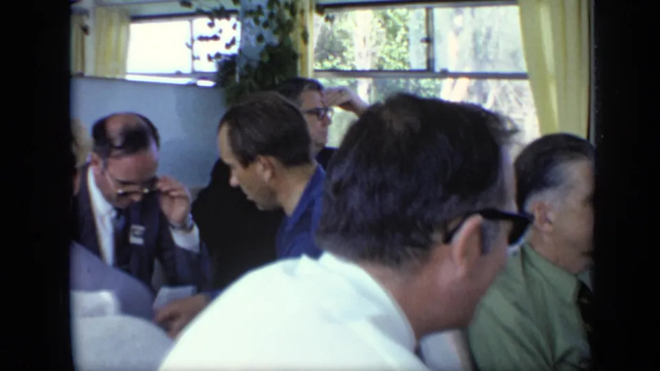 A crowded Moscow river boat cabin shows suited adult passengers, one adjusting glasses, with curtained windows and water beyond.