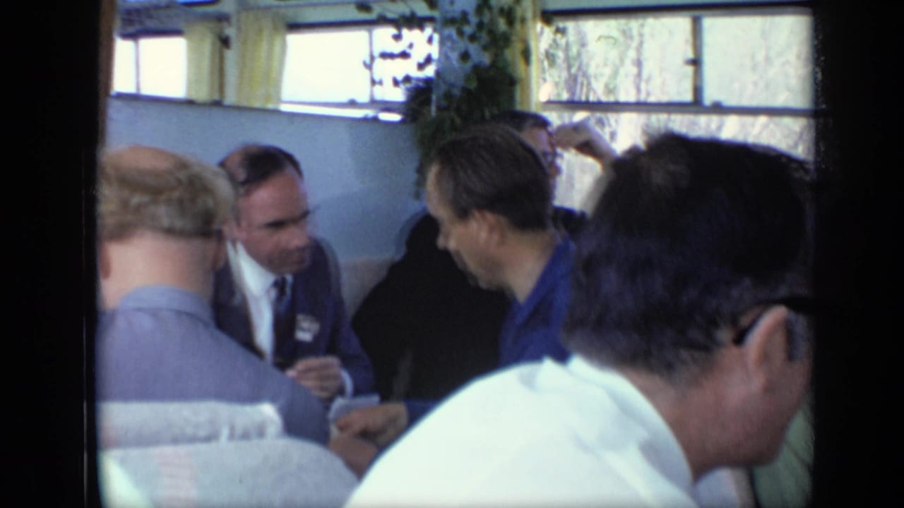 Men seated closely in a train compartment, some wearing suits and ties.