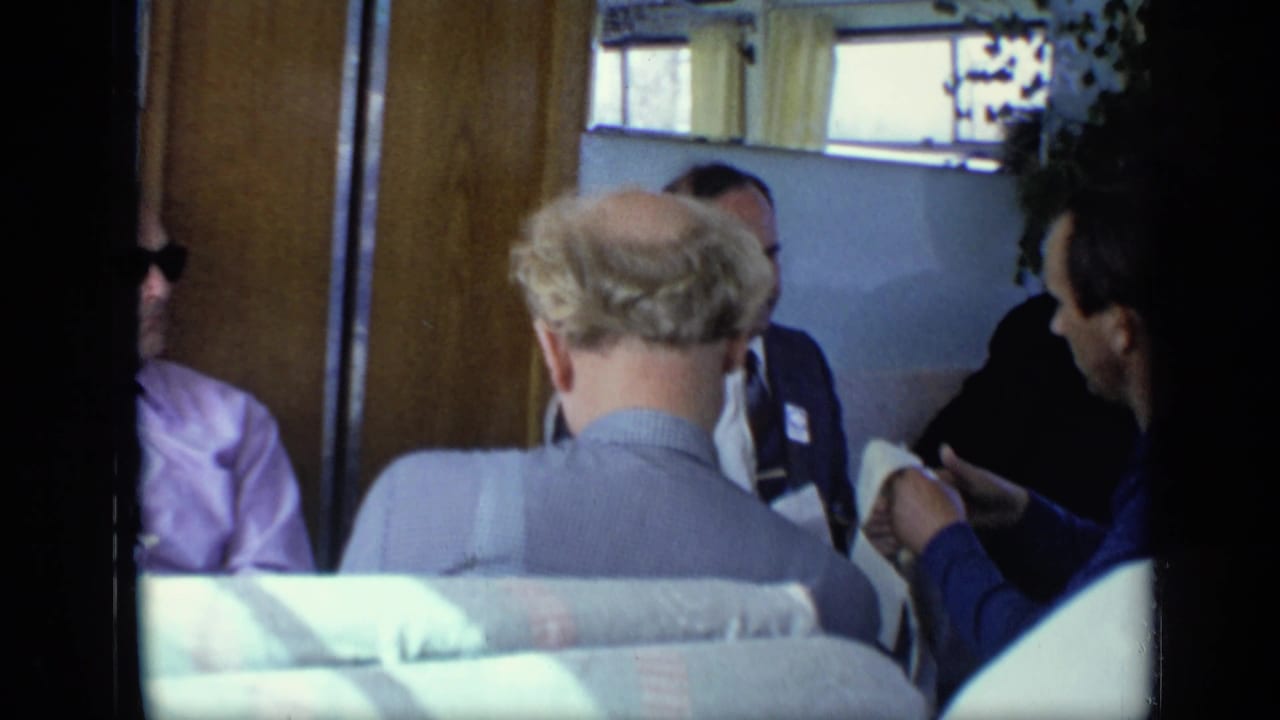Adult men continue a close conversation in a 1971 Moscow train interior, with seat backs, curtains, and wood trim visible.