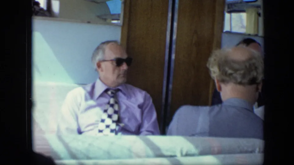 A man in sunglasses, light shirt, and checkered tie sits in the train compartment during the 1971 Moscow footage.