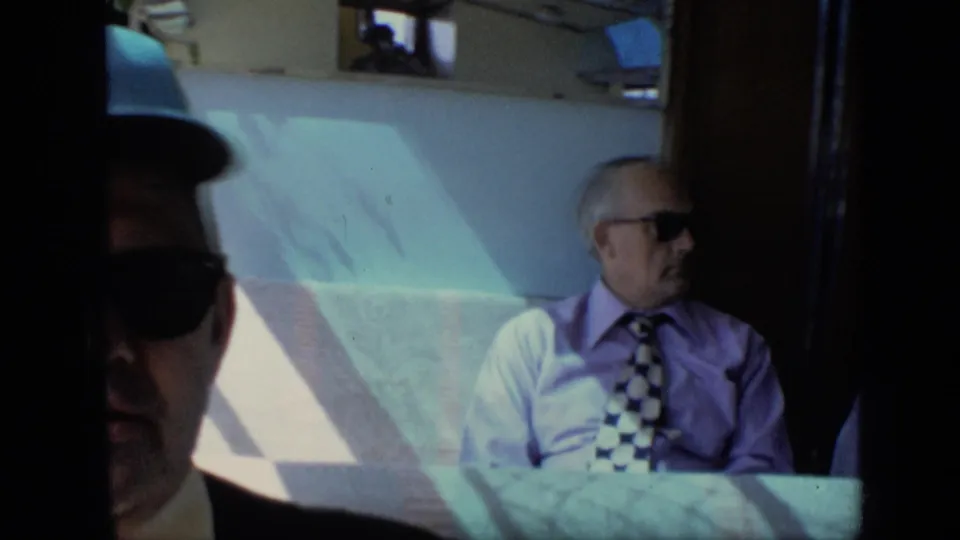 The 1971 Moscow train footage closes on a seated man in sunglasses and tie beside bright window light and pale upholstery.