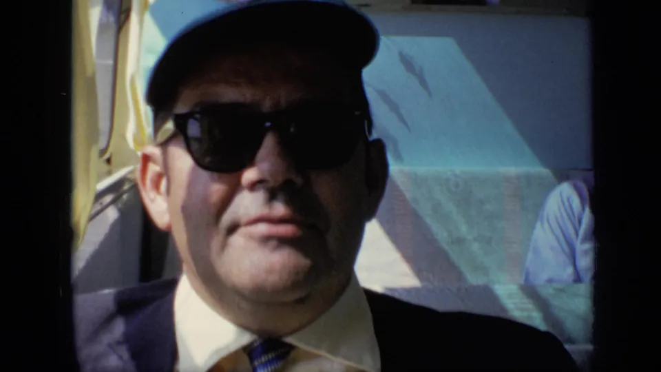 Adult man in a suit, tie, sunglasses, and light blue cap sits by a sunlit vehicle window in 1971 home-movie footage.