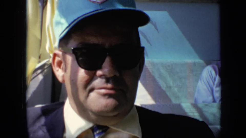 The same adult passenger sits close to a vehicle window, wearing dark sunglasses, a light blue cap, suit jacket, and tie.
