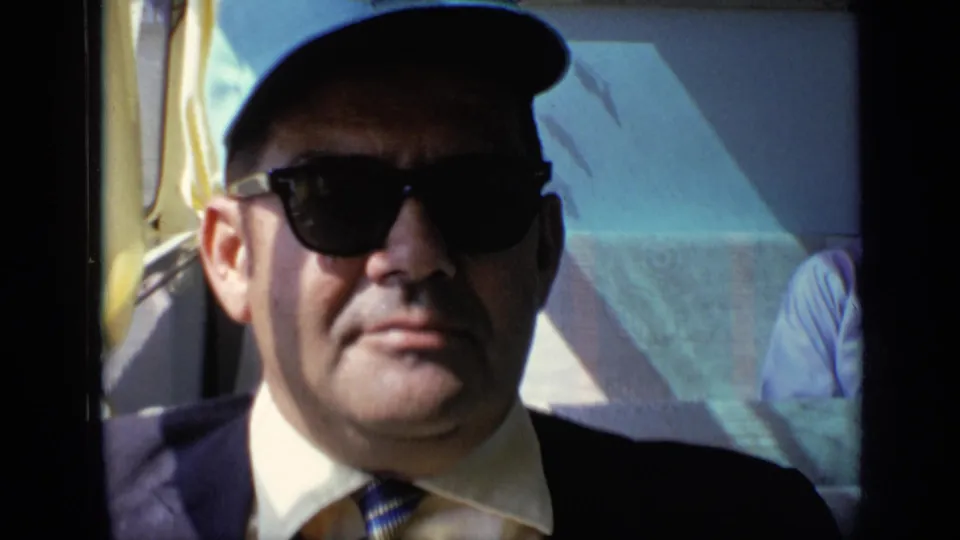 A close 8mm frame shows the adult man in sunglasses and cap seated by the window, with sunlight and interior trim behind him.