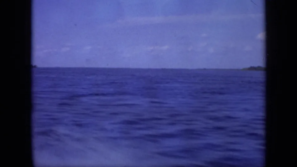 Soft 8mm frame of rippled water and a thin distant shore, continuing the boat approach view.