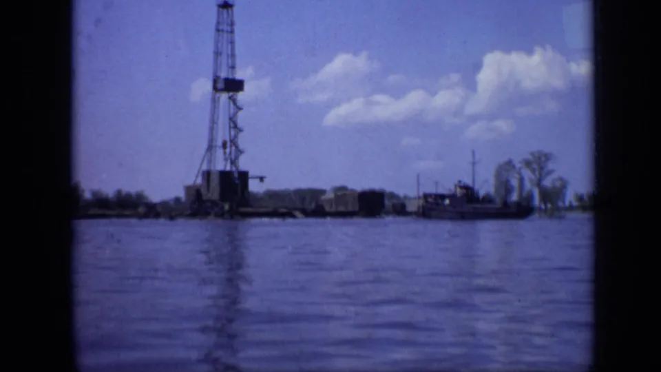 1971 8mm harbor view showing a tall industrial rig or tower, dockside structures, and a small boat on calm water.