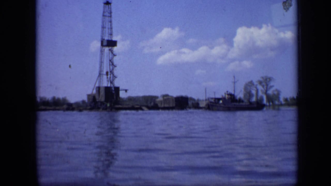 Tall oil derrick beside water with a small boat and cloudy blue sky.