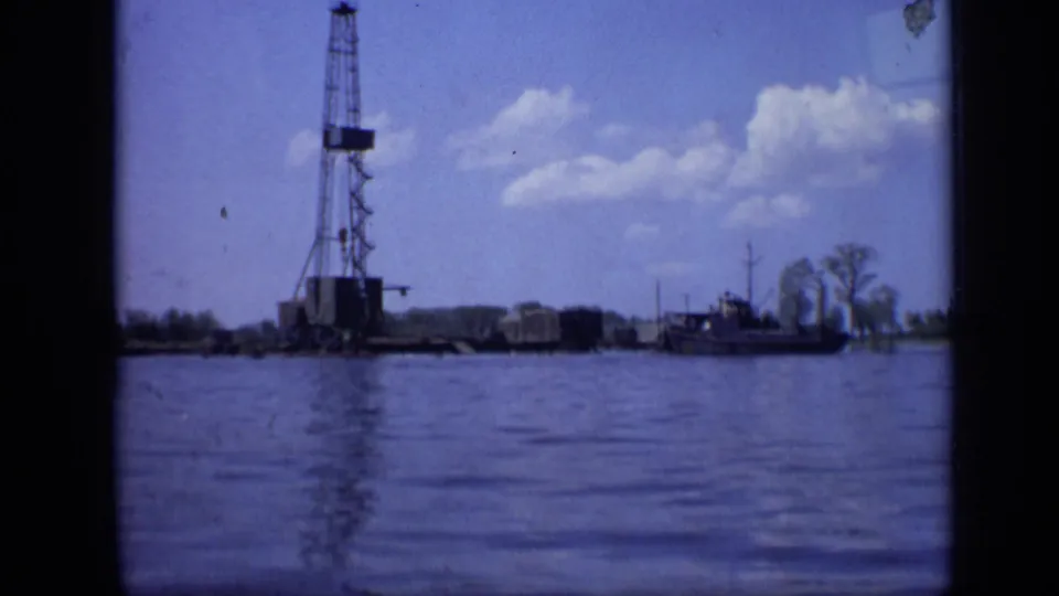 1971 Moscow home-movie frame showing an oil derrick on a shoreline, calm water in front, and a small boat nearby.