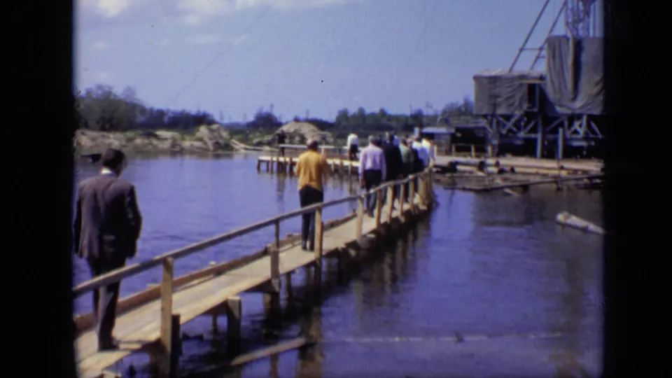 1971 Moscow 8mm frame showing adults on a wooden footbridge leading across calm water toward cranes and a covered structure.