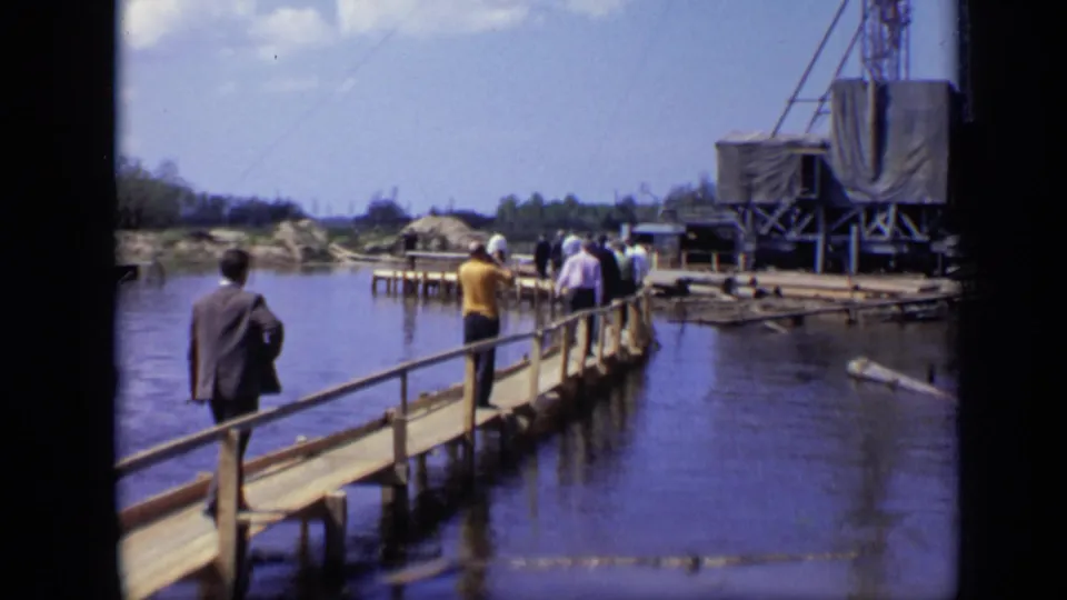 1971 Moscow 8mm frame of men walking over water on a wooden bridge toward an industrial waterside area under a bright sky.