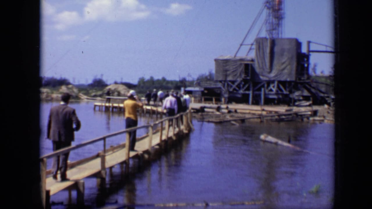 People walk along a wooden pier toward a tarped drilling rig over water.