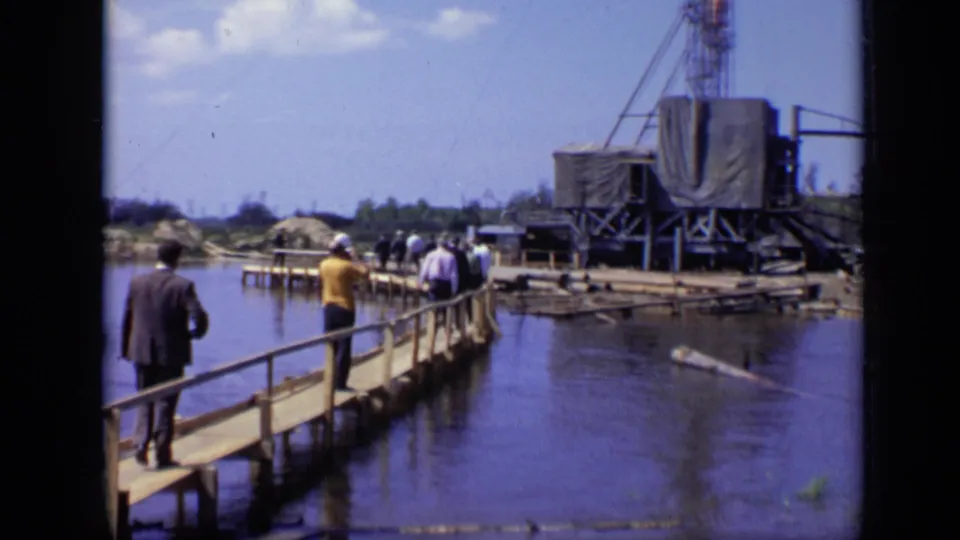 1971 Moscow home-movie frame with adults on a wooden pier approaching a tarped industrial drilling rig beside dark water.