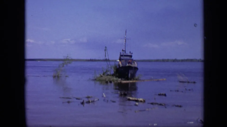 A dark working boat sits low on calm water in the 1971 Moscow river scene, with floating debris and a tree-lined far bank.