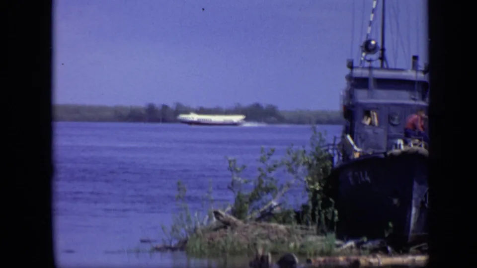 A white hydrofoil moves across the Moscow river in the distance while a dark boat and shoreline vegetation frame the foreground.