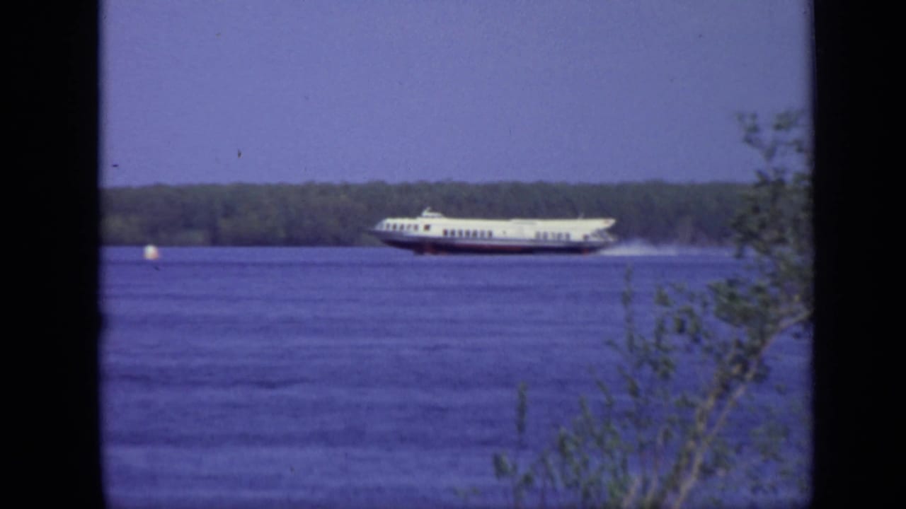 White ferry speeding across a river with spray behind it and trees on the far bank.