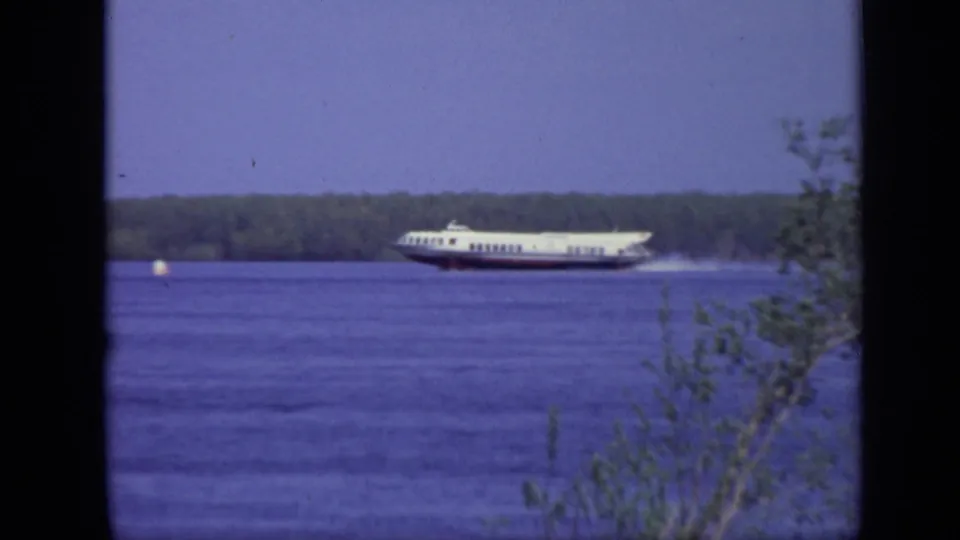 1971 Moscow frame of a white passenger ferry moving left to right across a wide river, leaving a pale wake near the wooded shore.