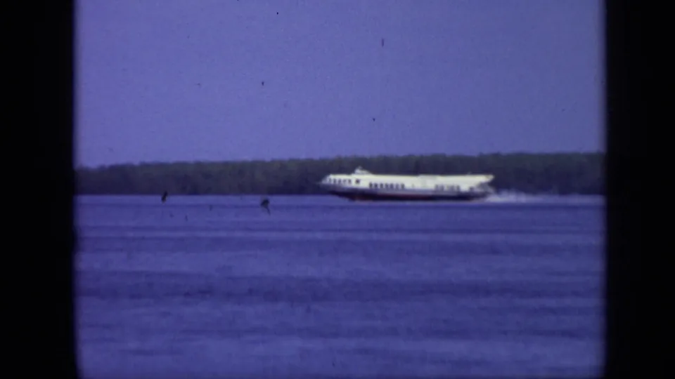 1971 Moscow home-movie frame showing a white passenger ferry moving across a broad river with a low wooded shoreline behind it.