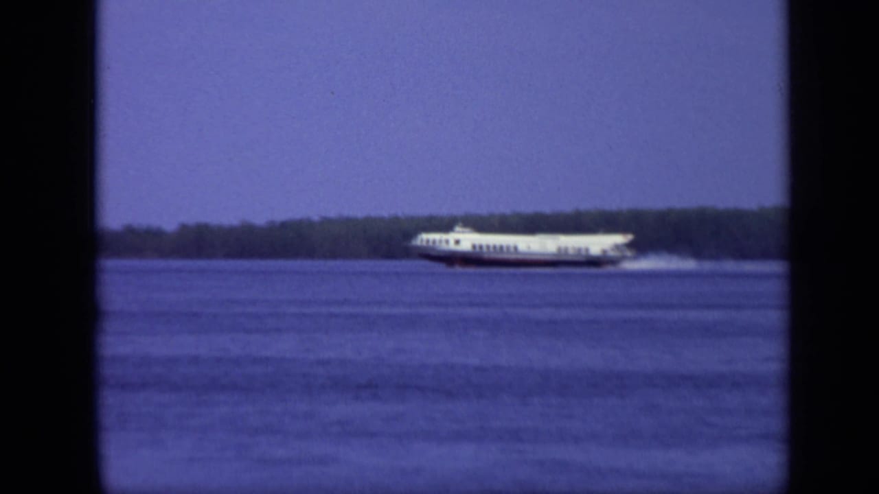 The passenger ferry continues across the Moscow river scene, framed through dark film edges with the wooded shoreline in the distance.