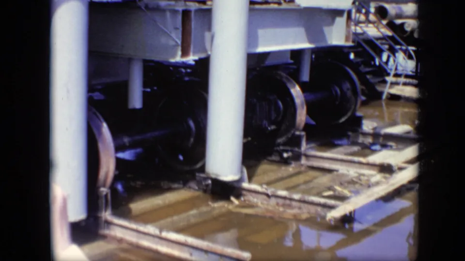 Close 8mm view of industrial ferry dock machinery, showing metal wheels, vertical pipes, beams, rails, and pooled water below.