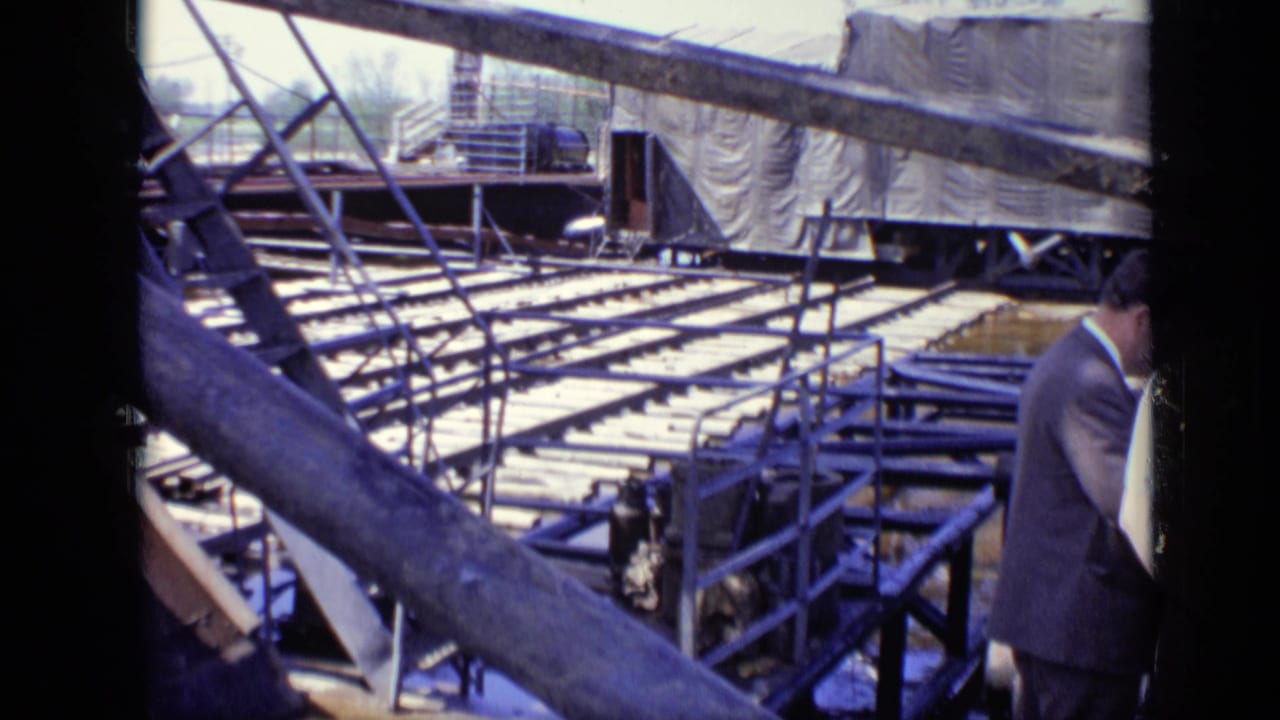 1971 Moscow footage frame with industrial framing, rows of planks, tarped equipment, and a partially visible suited visitor at the right edge.
