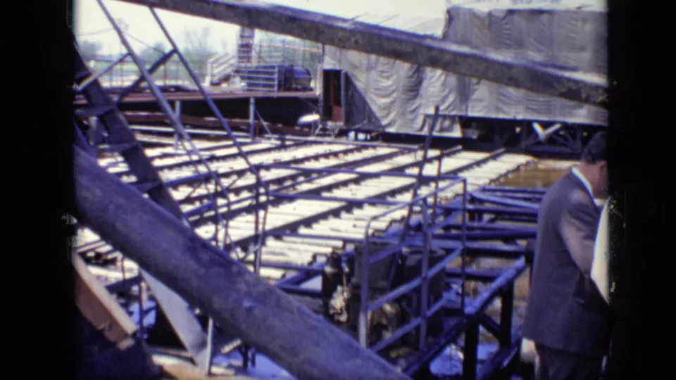1971 Moscow footage frame with industrial framing, rows of planks, tarped equipment, and a partially visible suited visitor at the right edge.