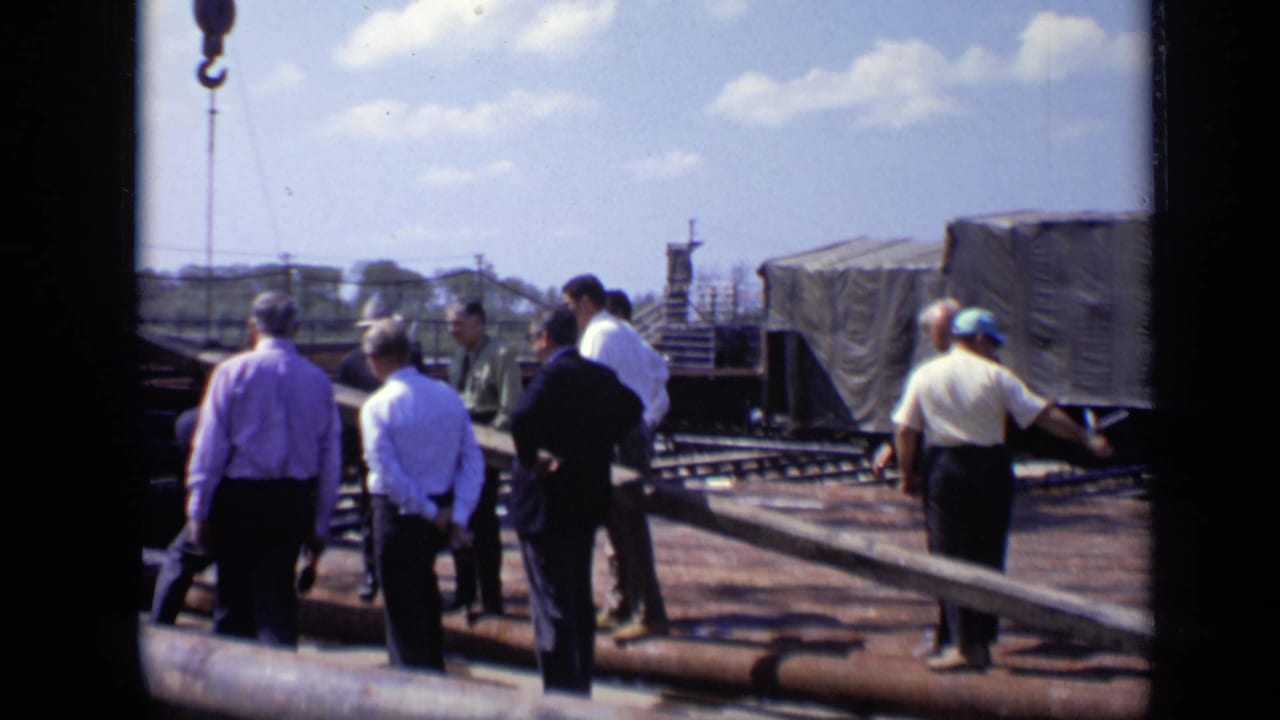 In 1971 Moscow footage, a group of men stands by rail tracks, beams, and covered freight cars at an outdoor industrial site.