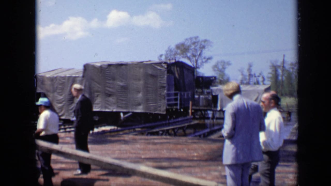 Men stand near a covered structure, pipes, and a construction area under bright sky.