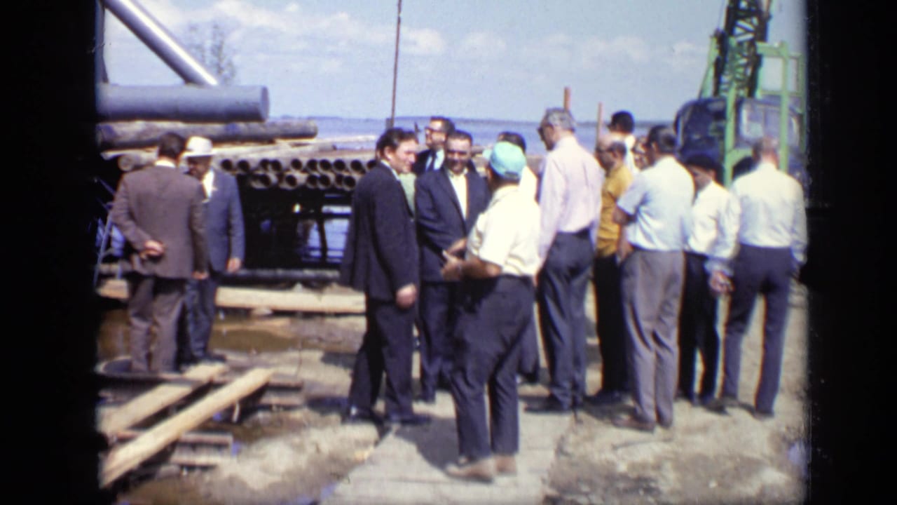 Men in suits, shirts, and hard hats appear to inspect stacked pipes and planks at a 1971 Moscow construction site near water.