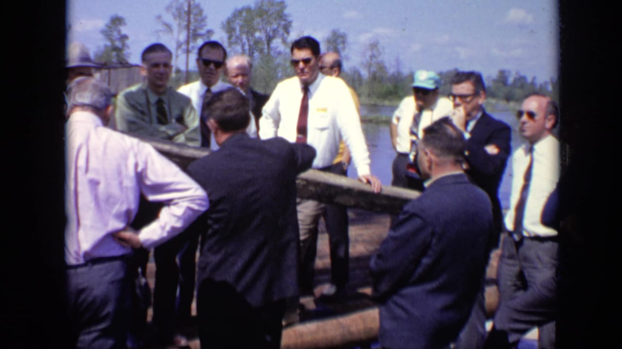 Men stand on a log raft beside river water while one man gestures during a group discussion.