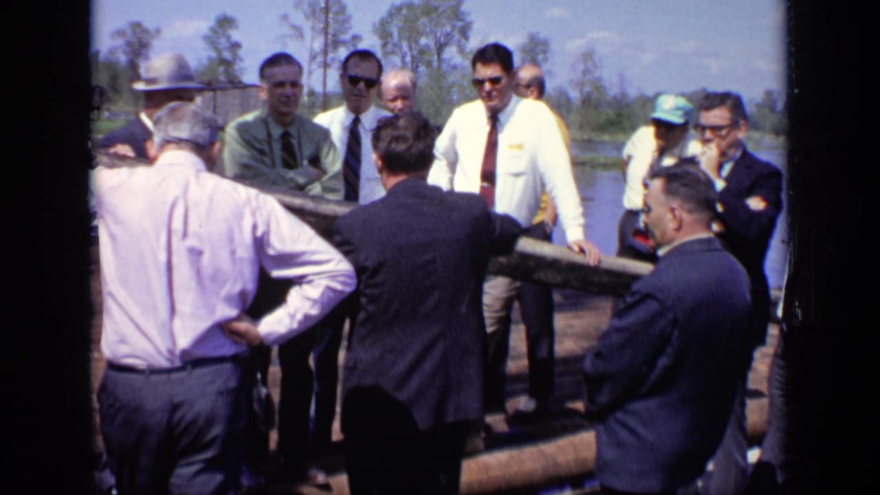 Adult men on a Moscow river log raft appear to listen to a speaker, with suits, sunglasses, and trees visible in daylight.
