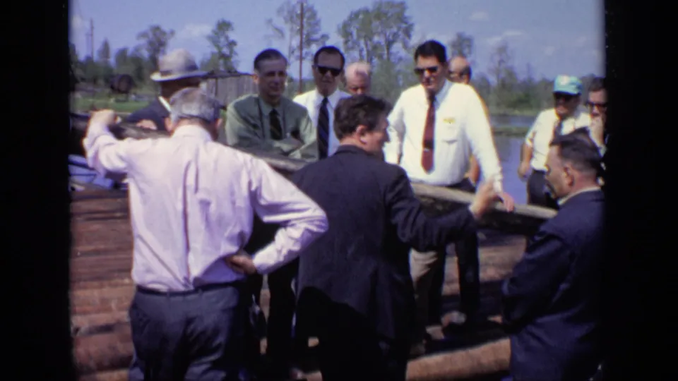 A handheld 8mm frame shows men gathered on a log raft in Moscow, with one man gesturing during the conversation.