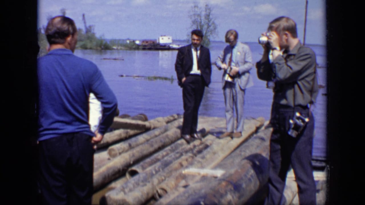 Men stand on logs beside open water while another man photographs them.
