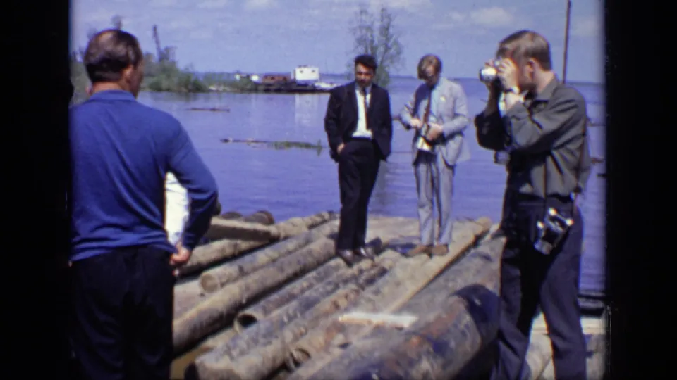 1971 Moscow home-movie frame showing men in suits on logs near water as a man with a camera photographs the group.