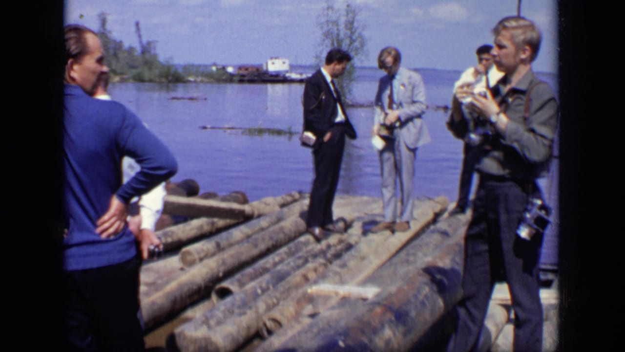 A photographer stands near suited men on logs at the waterfront.