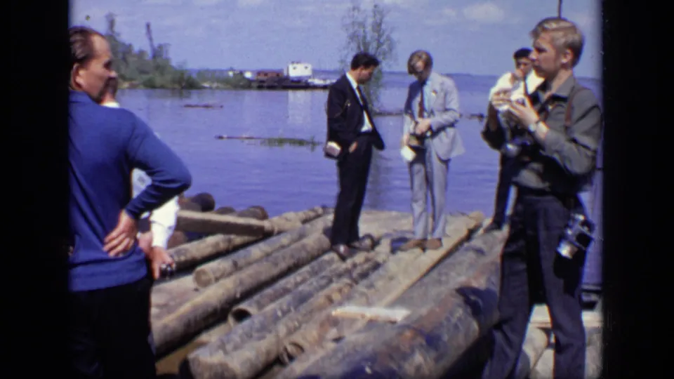 1971 Moscow waterfront frame with a photographer at right and suited men standing on logs beside calm water.