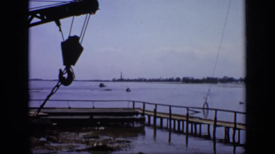 1971 Moscow riverside view with a dark crane hook in the foreground and a low wooden pier crossing calm water.