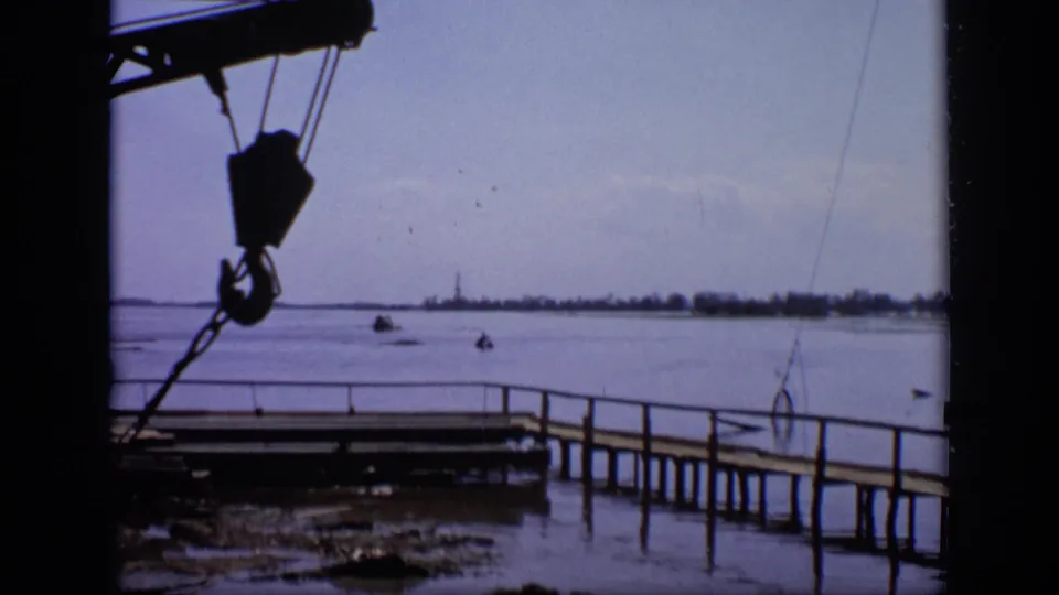 1971 Moscow home-movie frame showing a crane hook over a pier and open water with a distant low shoreline.
