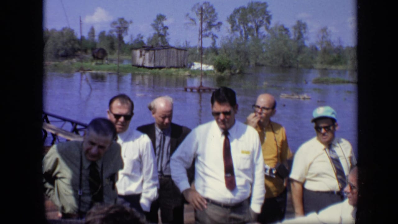 Adult men in shirts, ties, and sunglasses stand near floodwater and a small shed in 1971 home-movie footage from Moscow.
