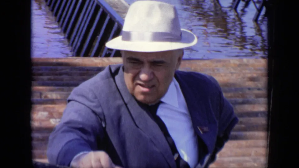 1971 Moscow home-movie frame showing an adult man in a white fedora, blue suit, and dark tie leaning forward near water.