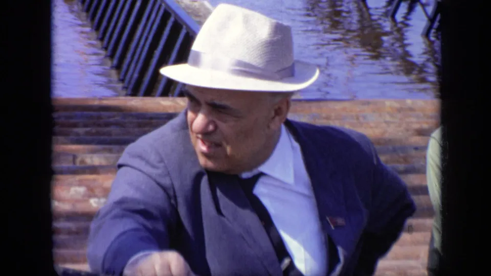 1971 Moscow home-movie frame of a suited adult man in a white fedora looking sideways beside water and stepped stonework.